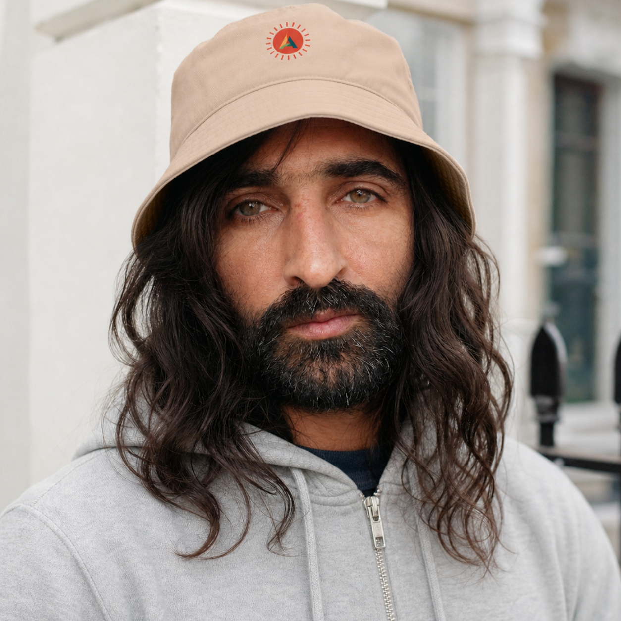 A male model wearing the Aether Peak organic cotton bucket hat with the Sunburst logo and a gray hoodie outdoors.