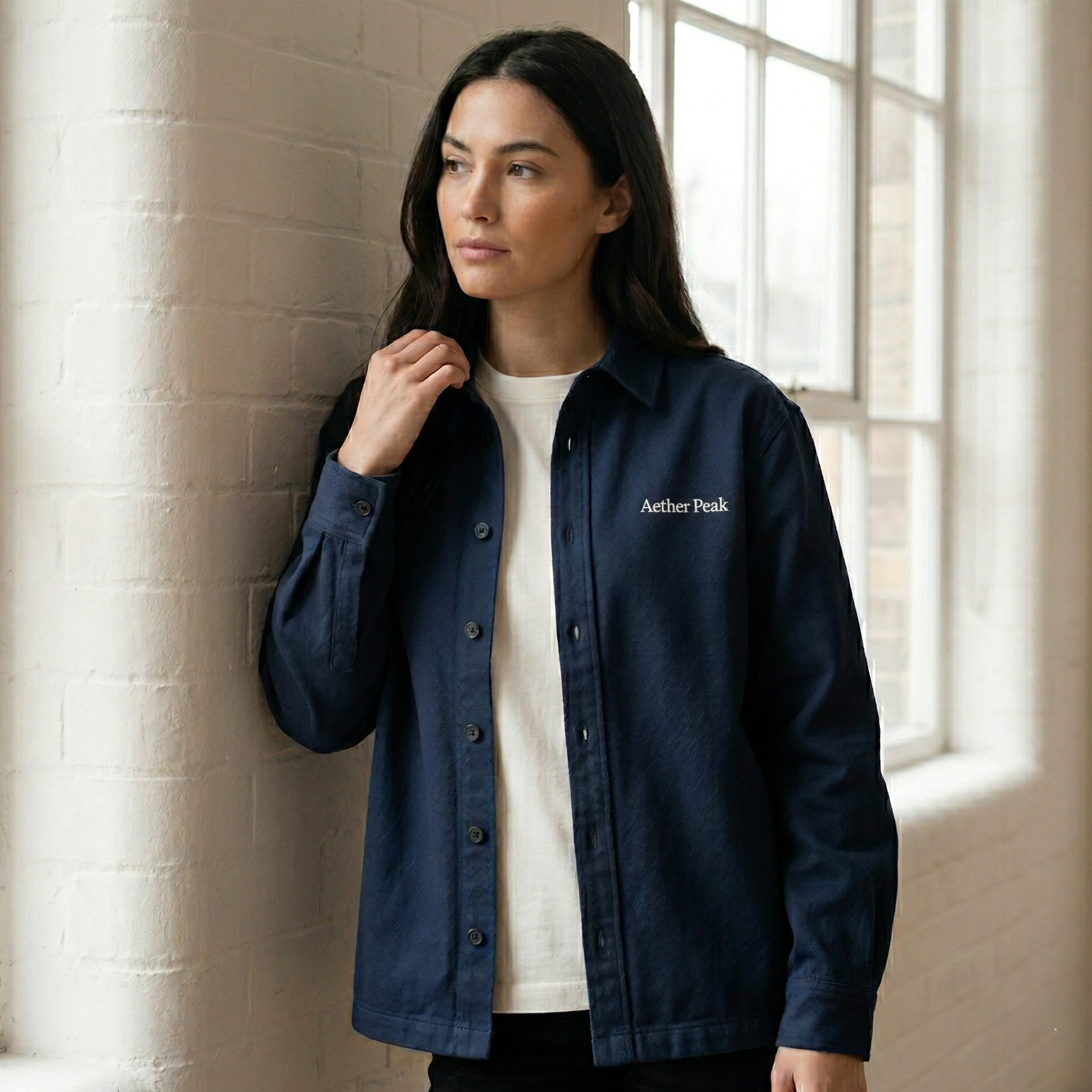Woman wearing a French Navy organic cotton twill overshirt with 'Aether Peak' logo against a light-colored wall.