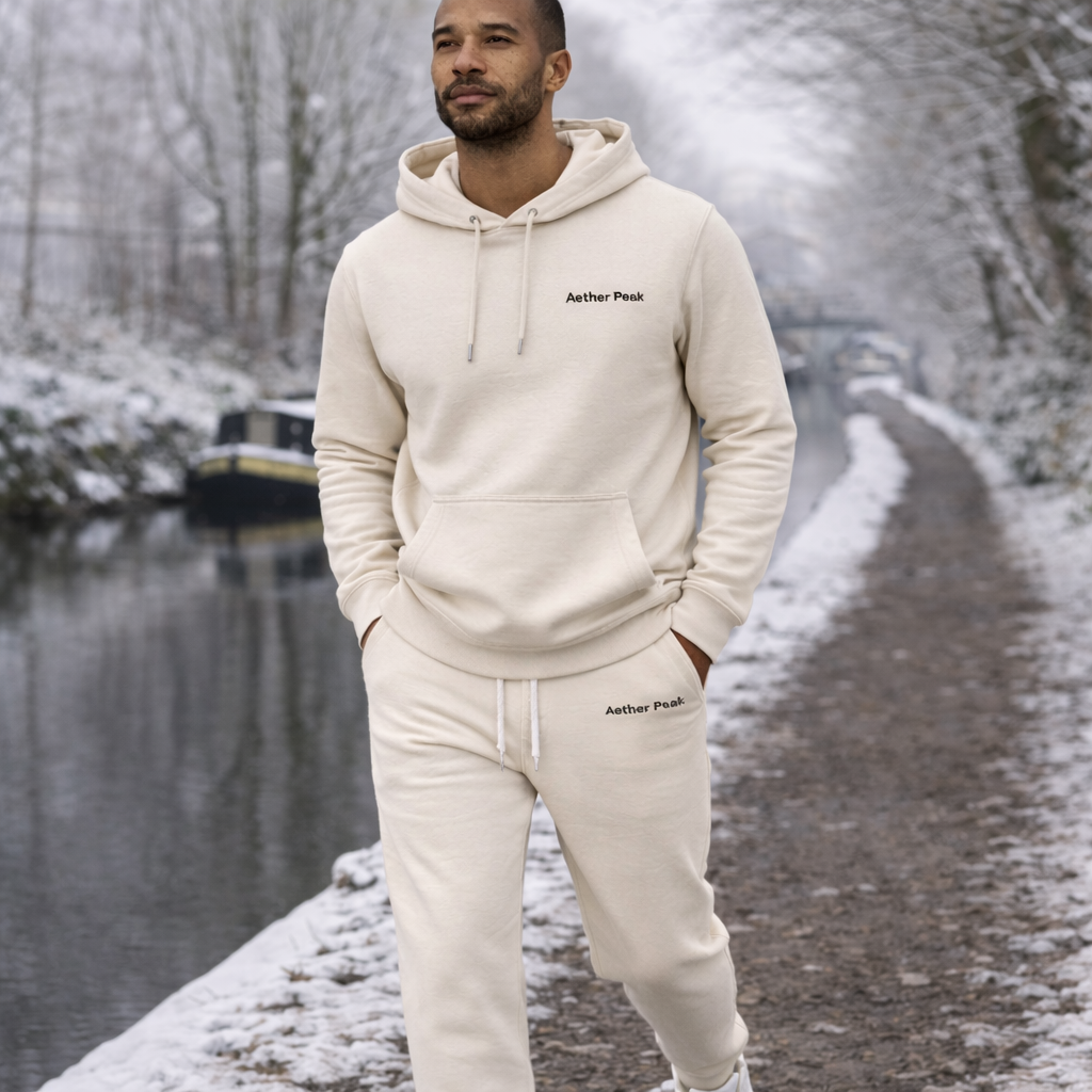 A man wearing a Desert Dust hoodie and joggers with Aether Peak embroidery, standing on a snowy path along a canal 