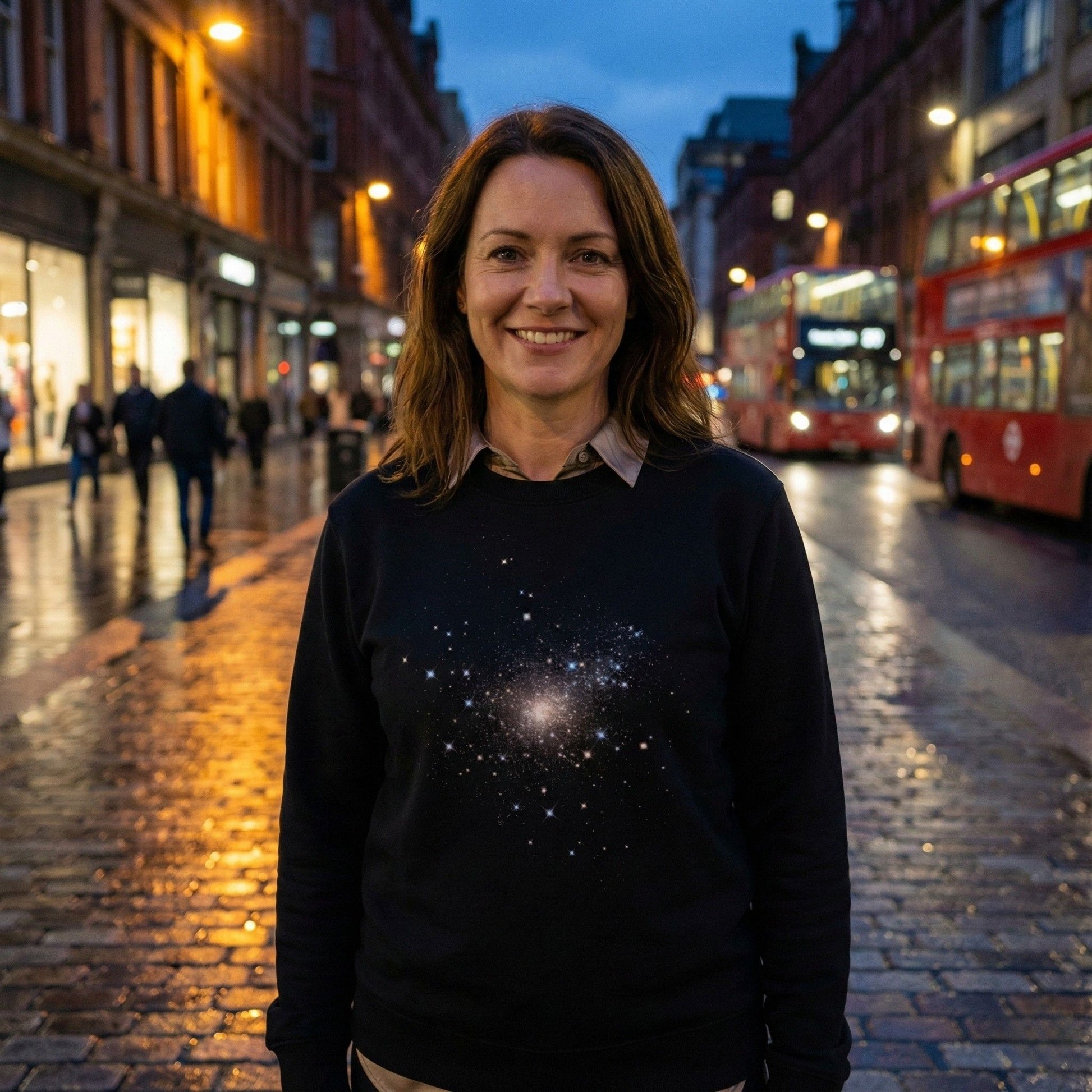 A woman wearing a black Aether Peak Starry Sky eco sweatshirt with a galaxy design, standing on a wet city street at dusk with red buses and shop lights in the background.
