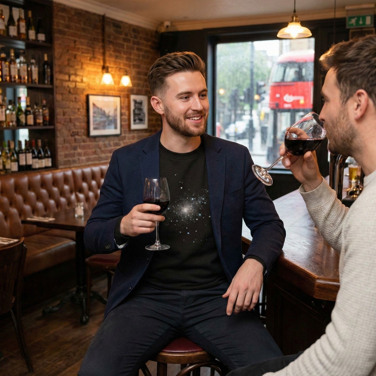 Man wearing a black Aether Peak Starry Sky eco sweatshirt and navy blazer, sitting in a cosy bar with exposed brick, holding a glass of red wine and talking with a friend, with a red double-decker bus visible through the window.
