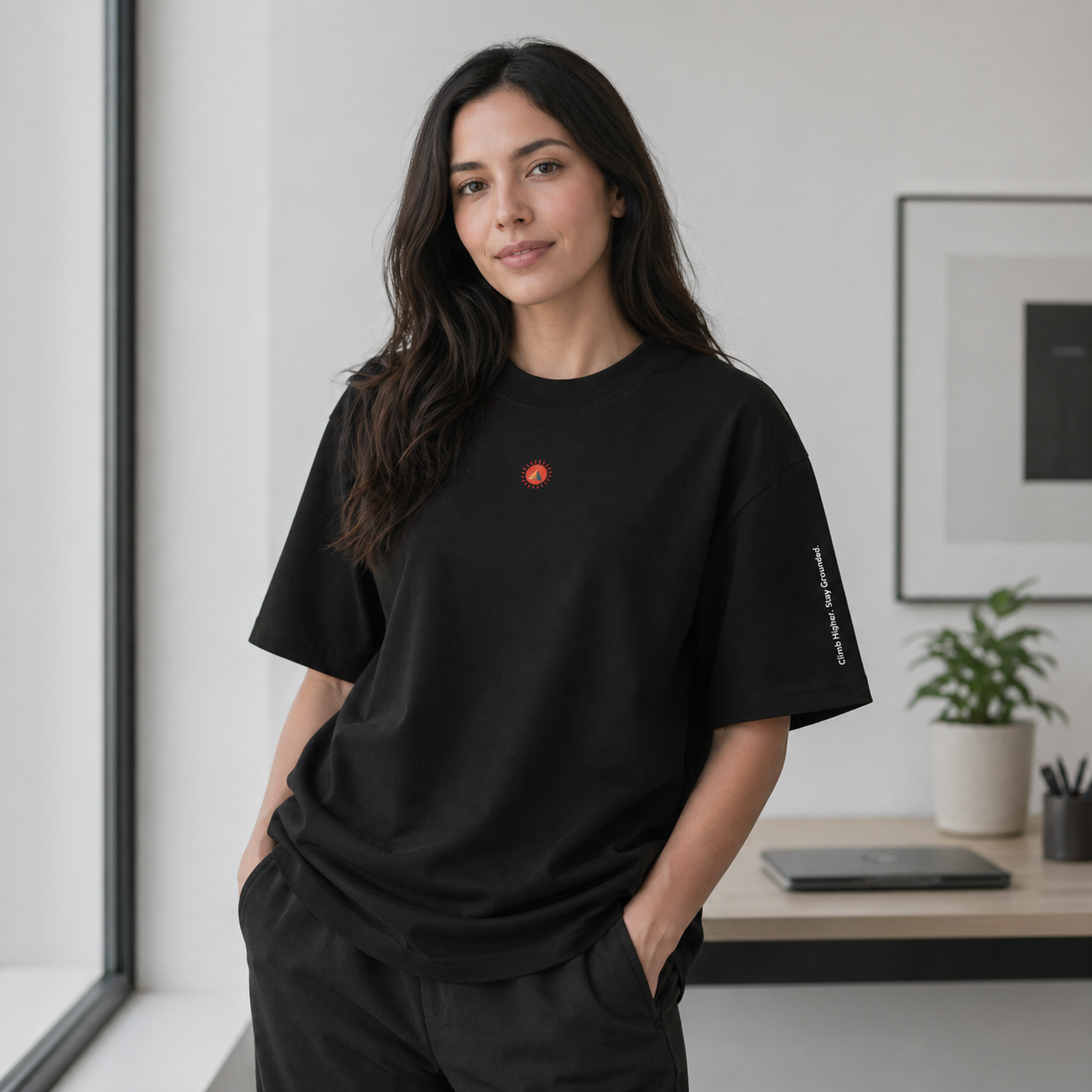  A female model wearing the black oversized organic cotton tee with subtle sunburst chest logo and sleeve text, styled in a minimalist indoor setting.