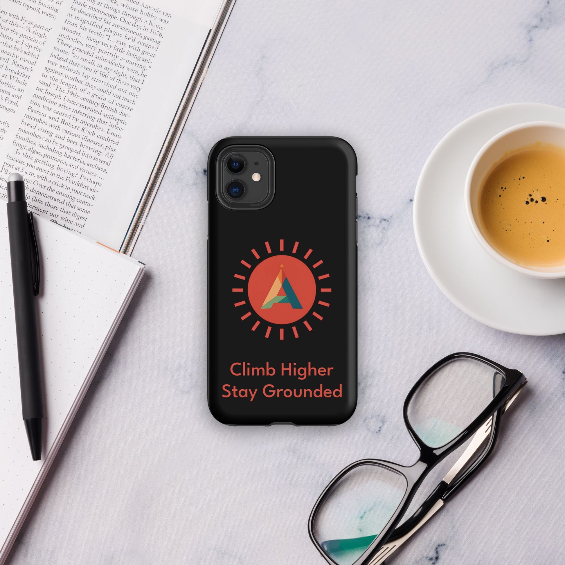 Phone case with the Aether Peak Sunburst and Climb Higher Stay Grounded quote on a marble surface with a book, pen, and coffee.