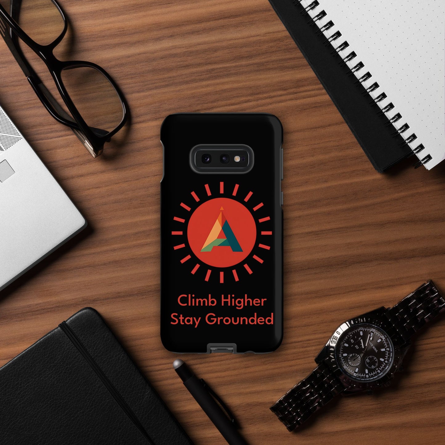 Phone case with the Aether Peak Sunburst logo and "Climb Higher.  Stay Grounded" text on a desk with office items
