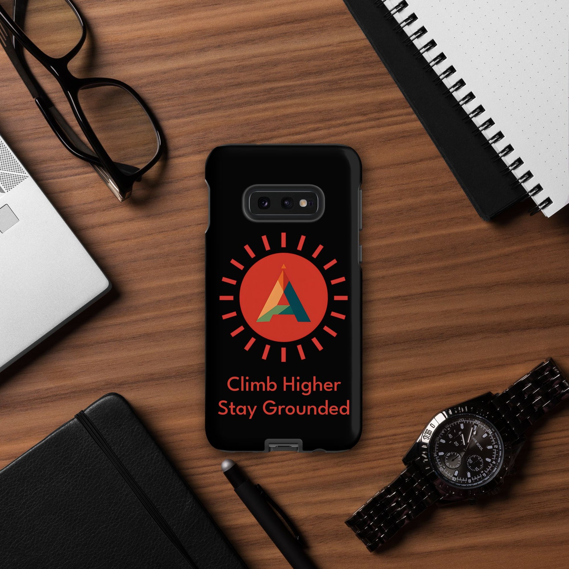 Phone case with the Aether Peak Sunburst logo and "Climb Higher.  Stay Grounded" text on a desk with office items