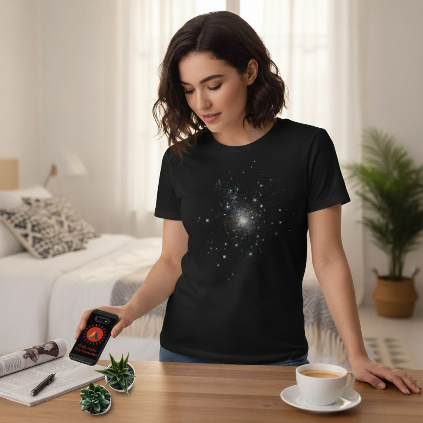 A woman wearing a black t-shirt with a starry sky design, holding a phone with the Aether Peak logo in a bedroom setting.