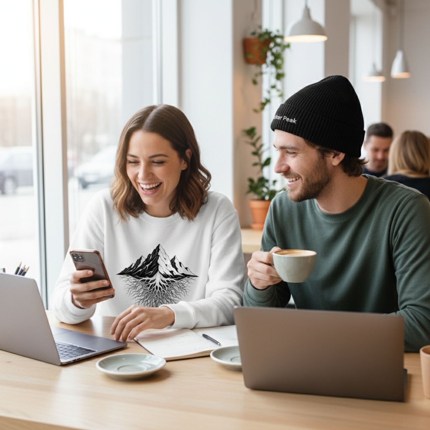 Smiling woman in a white Aether Peak Mountain Roots sweatshirt at a café, working with a friend with on 2 laptops at  a table.
