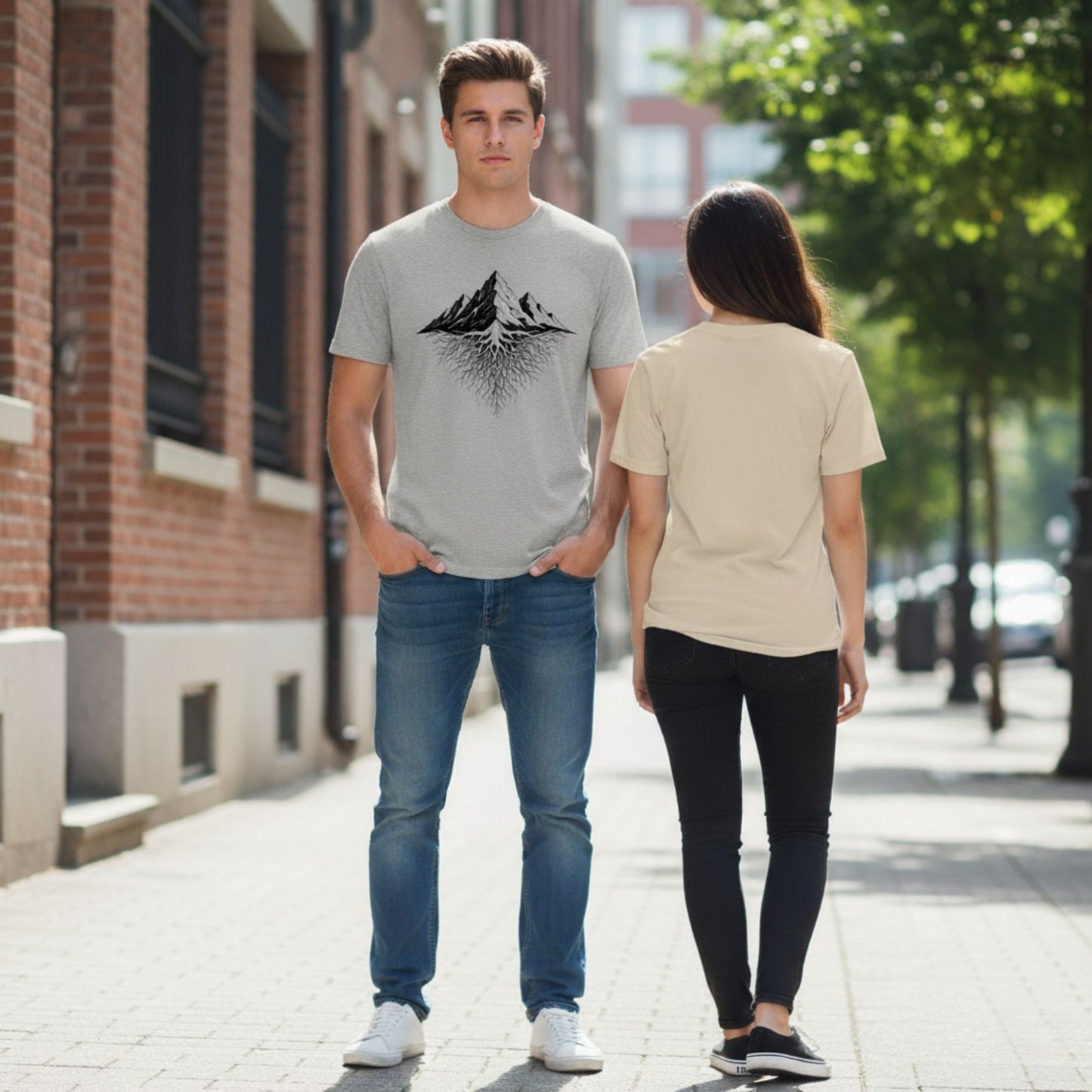 A man in the grey Aether Peak Roots mountain tee is standing with a woman in the desert dust tee on a sunny city sidewalk.