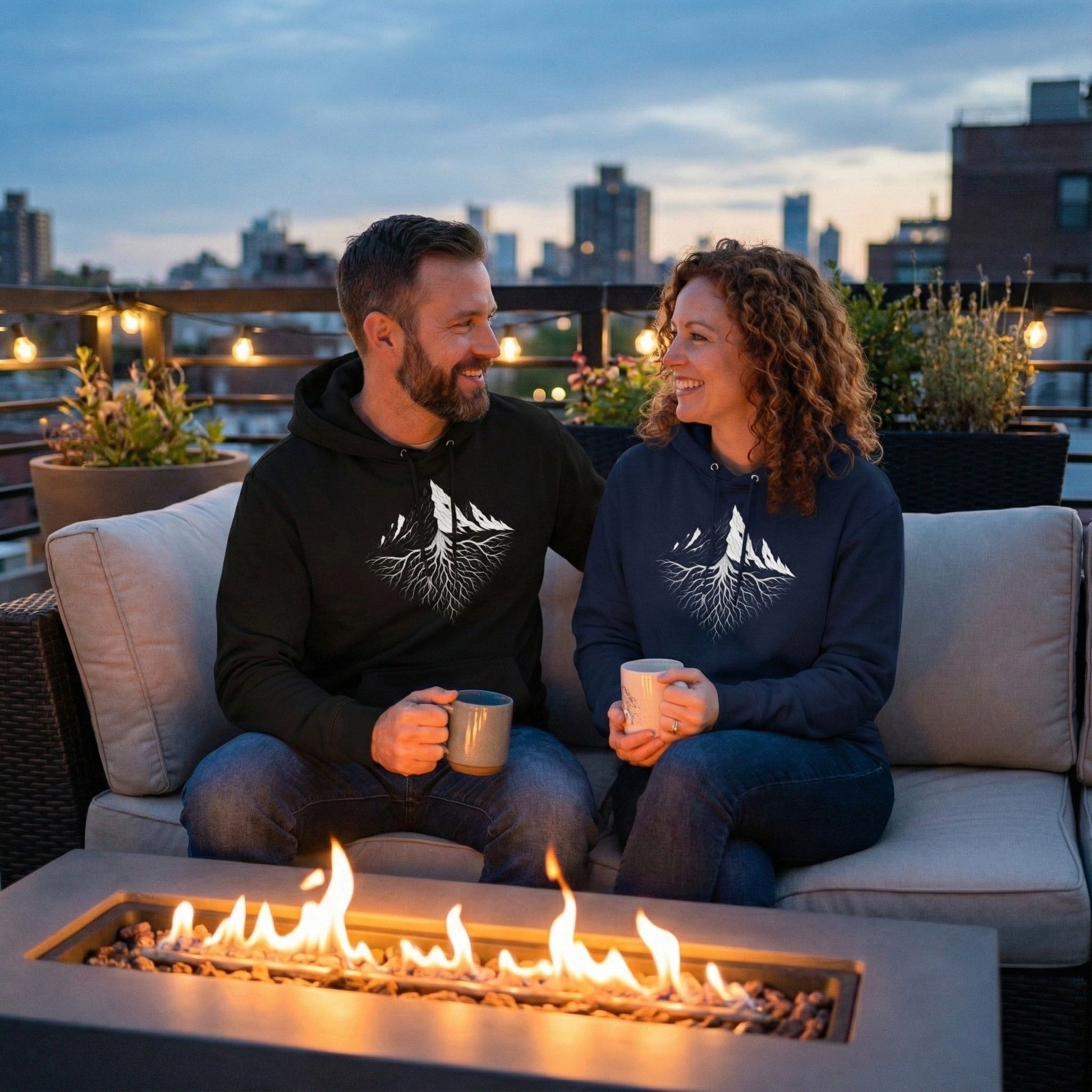 a man and woman sitting on a rooftop sofa at dusk, wearing Aether Peak Roots hoodies with white mountain and roots graphic, holding mugs in front of a modern gas fire pit.
