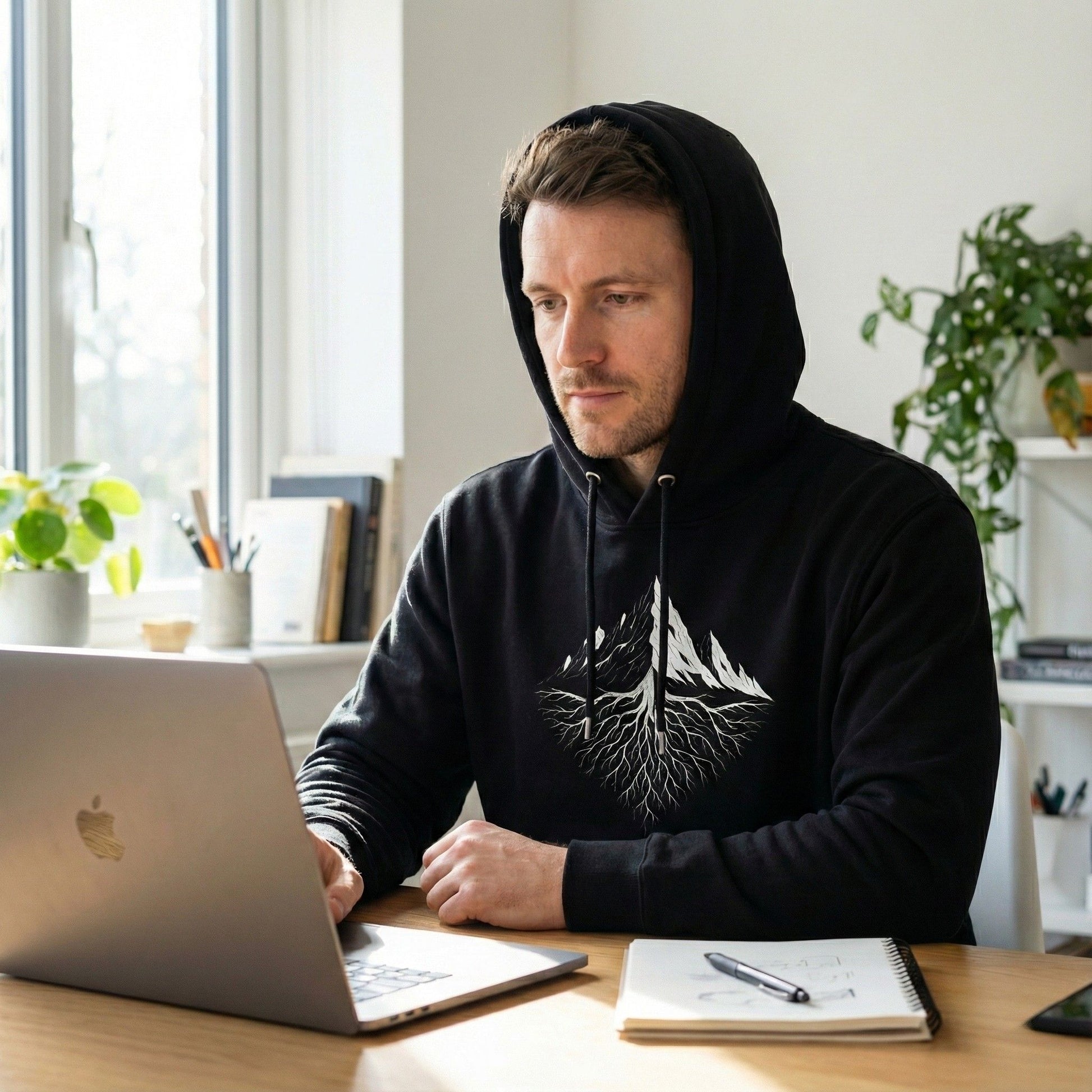 Man wearing a black hoodie with a white design, sitting at a desk with a laptop and notebook.