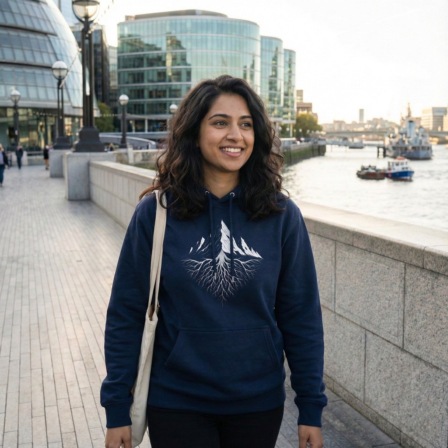 A woman wearing a French Navy blue hoodie with a white Mountain Roots graphic, standing by a waterfront with modern buildings in the background.