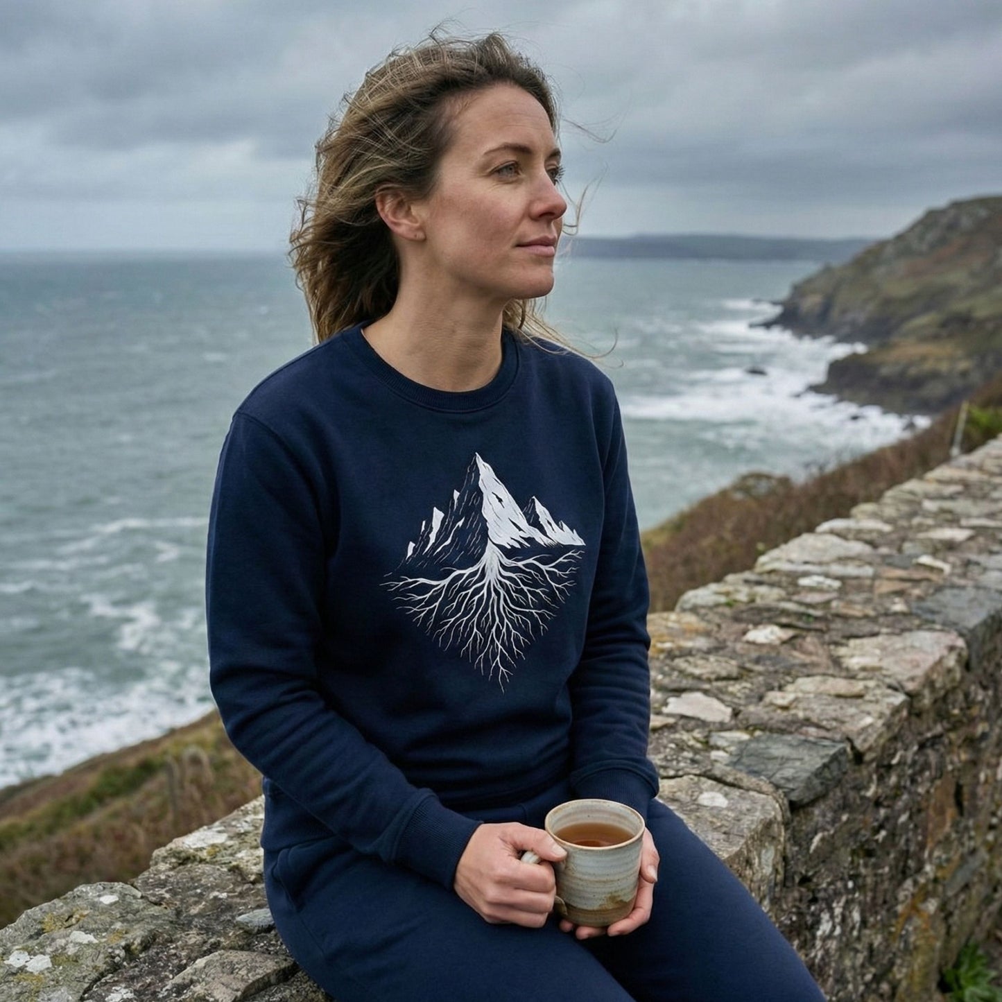 Woman wearing a French navy Aether Peak Roots sweatshirt, sitting on a stone wall by the sea and holding a mug on a windy coastal day.
