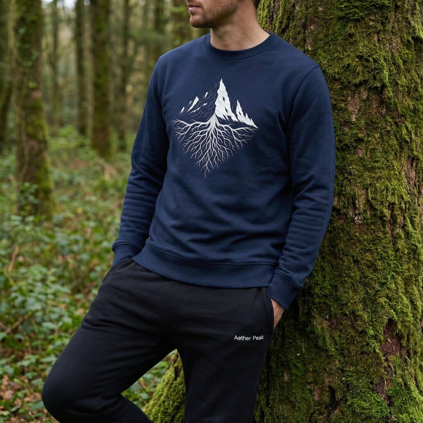 A man wearing a French navy Aether Peak Roots sweatshirt, leaning against a moss-covered tree in a quiet forest.
