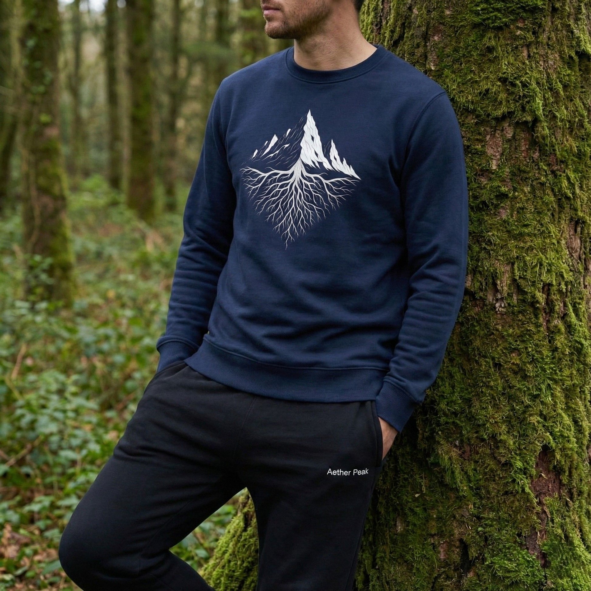 A man wearing a French navy Aether Peak Roots sweatshirt, leaning against a moss-covered tree in a quiet forest.
