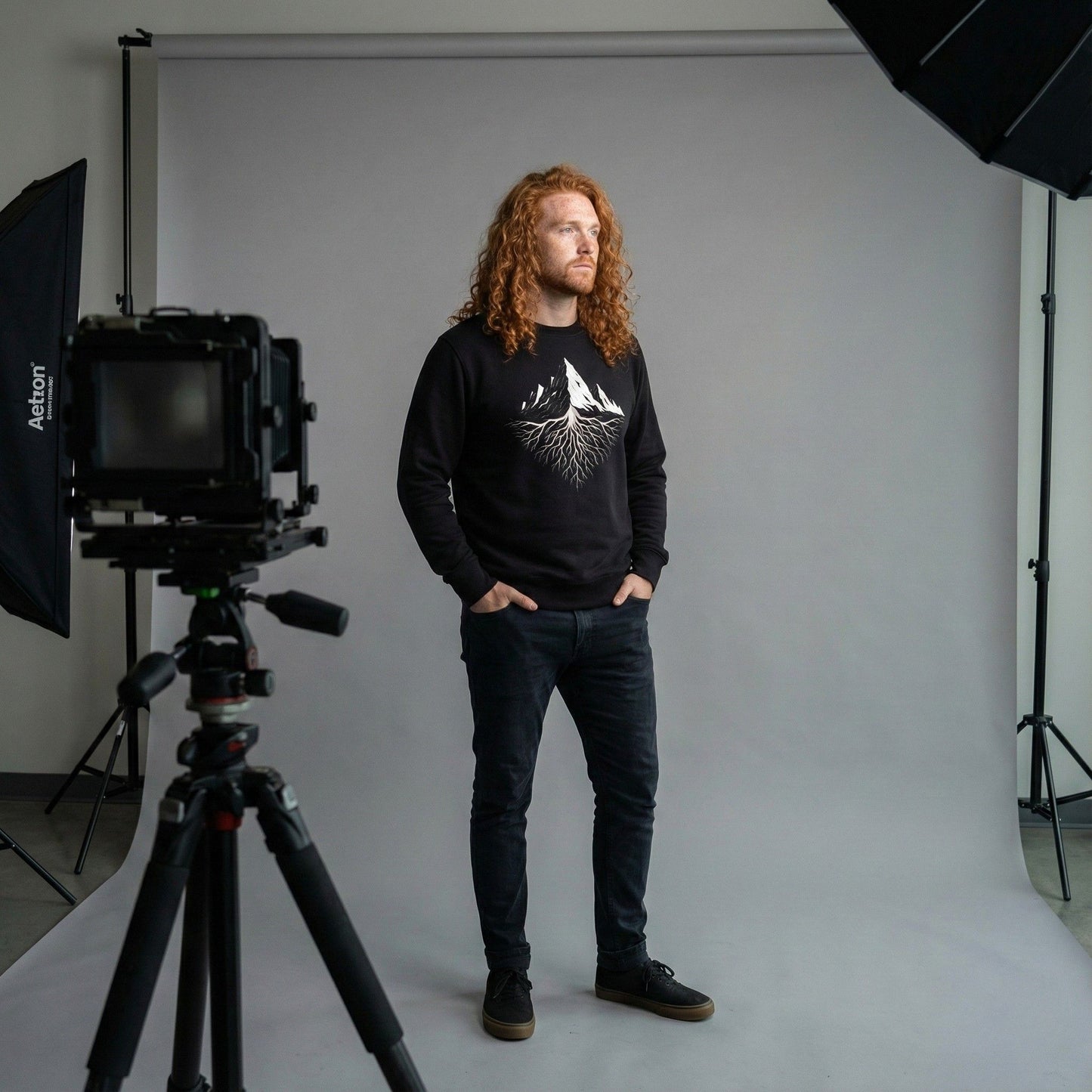 A man standing in a photo studio wearing a black Aether Peak sweatshirt with white mountain and roots graphic on the chest.
