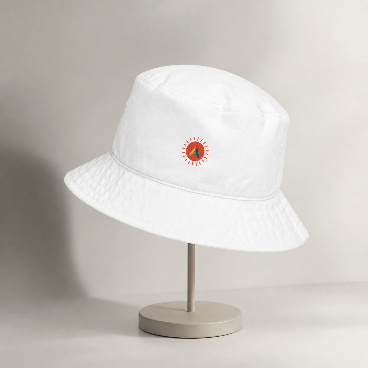 White Aether Peak organic cotton bucket hat with the Sunburst logo resting on a stand on an off-white background.