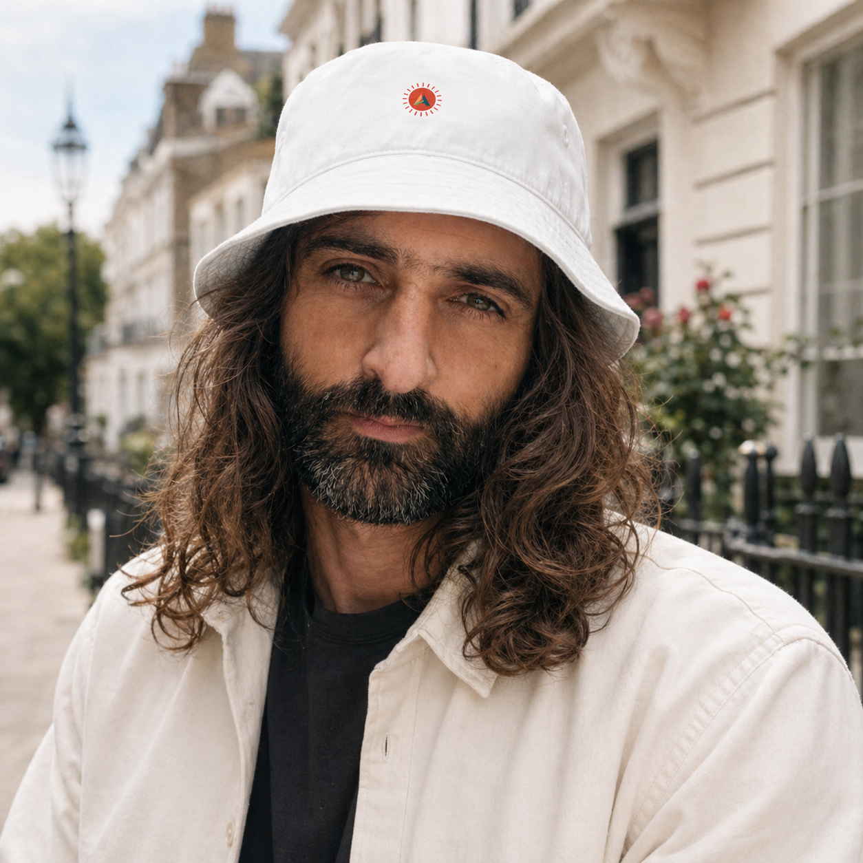 A male model with long hair and beard wearing a white Aether Peak Sunburst organic cotton bucket hat in an urban setting.
