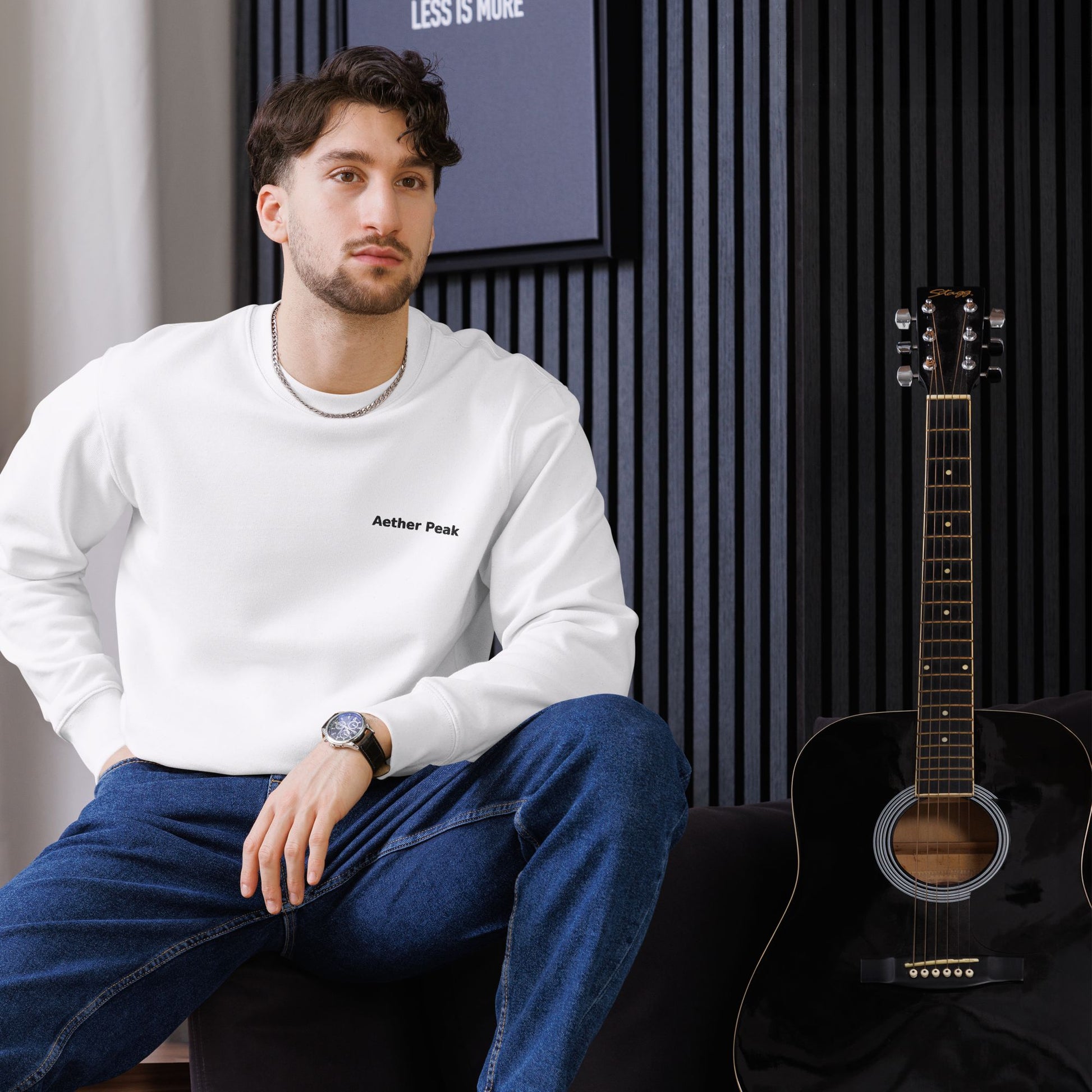 A man sitting next to an acoustic guitar wearing a white Aether Peak sweatshirt in a room with striped walls.