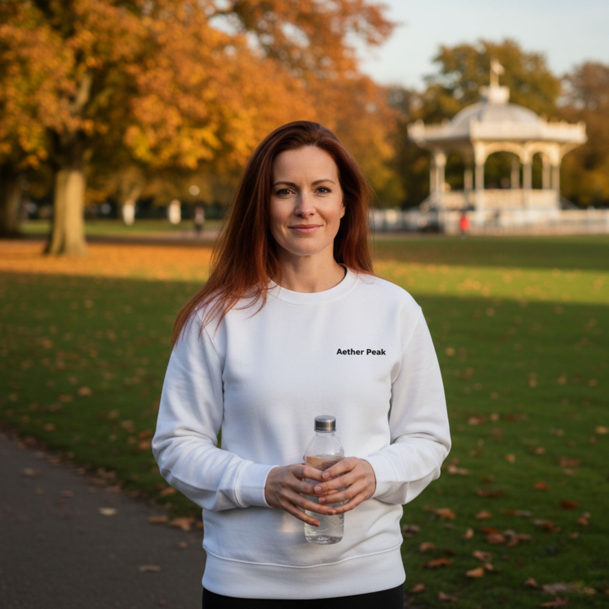 Woman in a white Aether Peak sweatshirt holding a water bottle in a park with autumn trees and a gazebo.