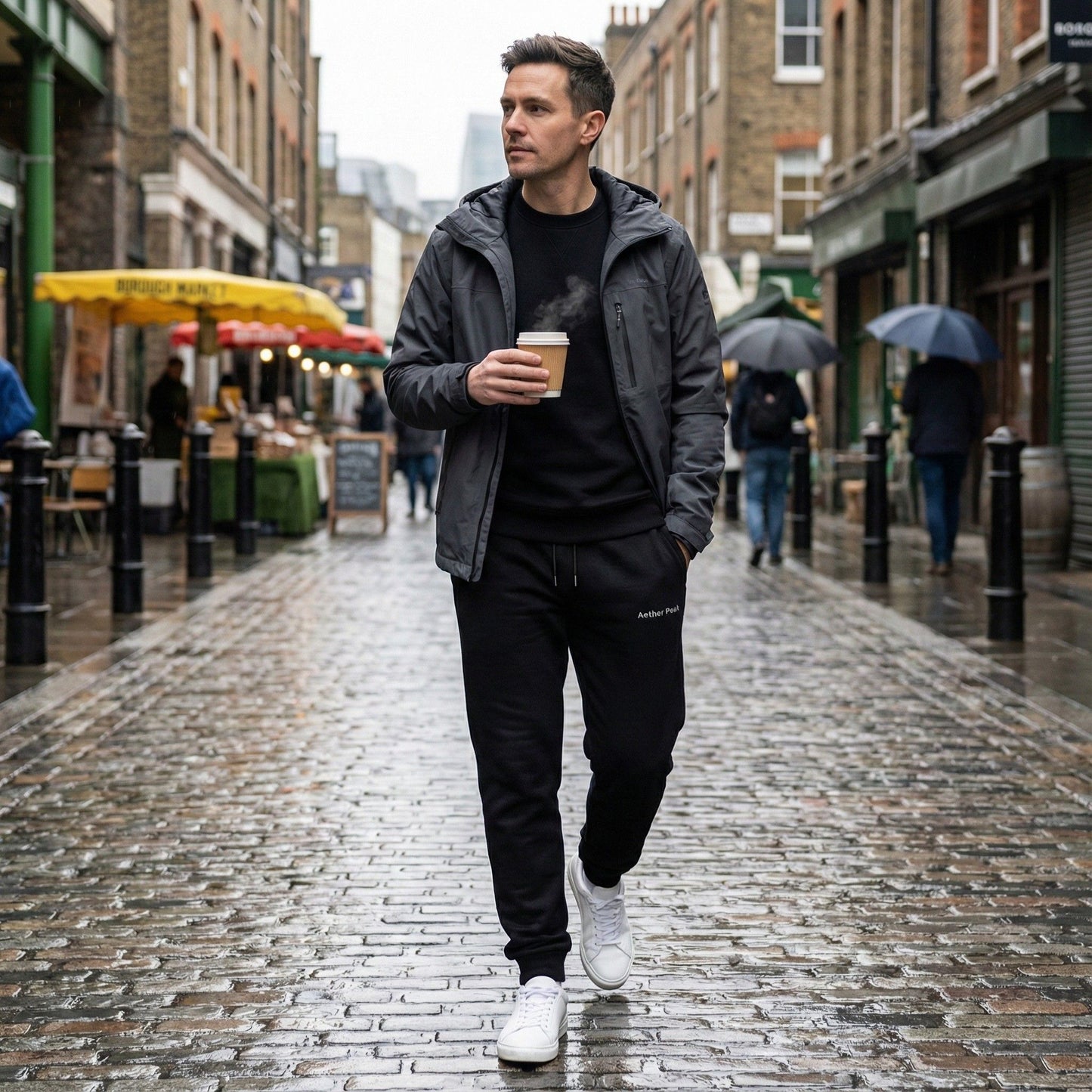 A man walking through a rainy city street in black Aether Peak The Zenith joggers, holding a takeaway coffee.