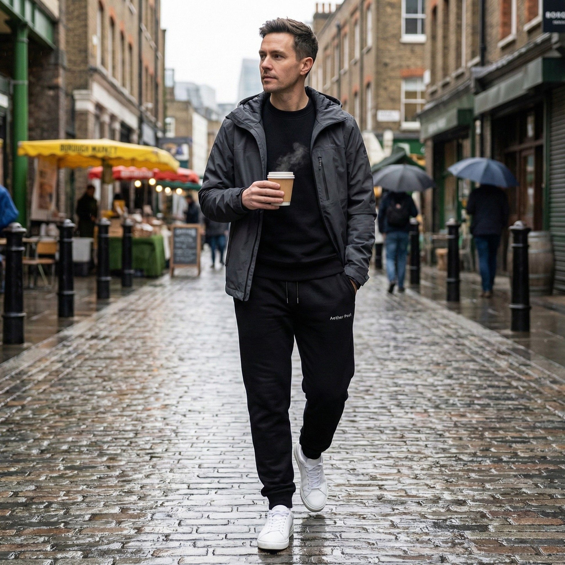 A man walking through a rainy city street in black Aether Peak The Zenith joggers, holding a takeaway coffee.