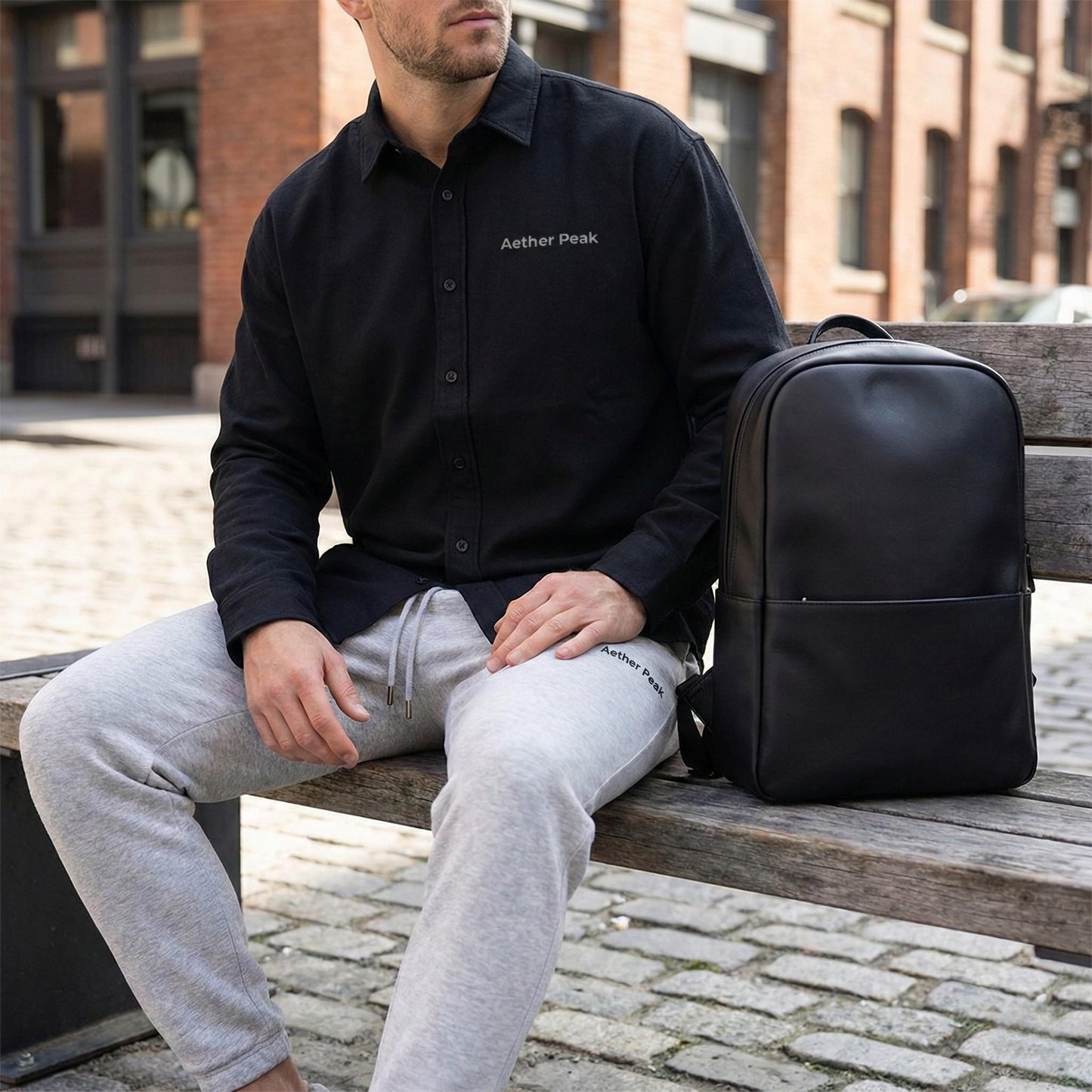 A man wearing a black Aether Peak organic cotton twill overshirt, sitting on a bench with a black backpack and Heather Grey Aether Peak organic joggers in an urban street setting.