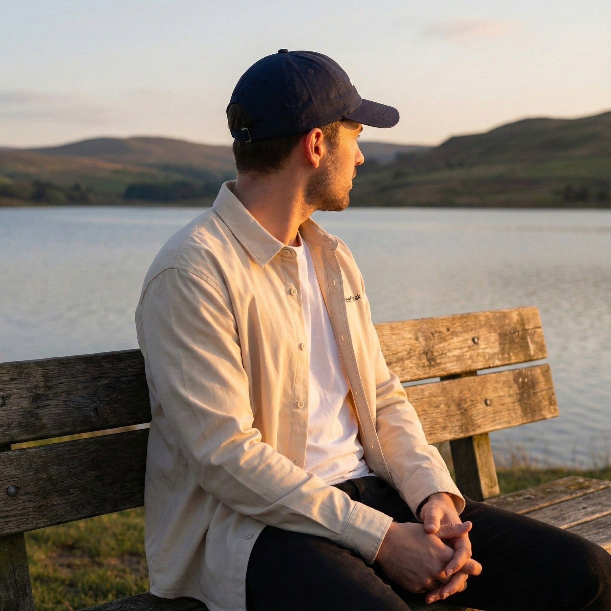 A man wearing a desert dust Aether Peak organic twill overshirt and French Navy Organic cap, sitting on a lakeside bench at sunset