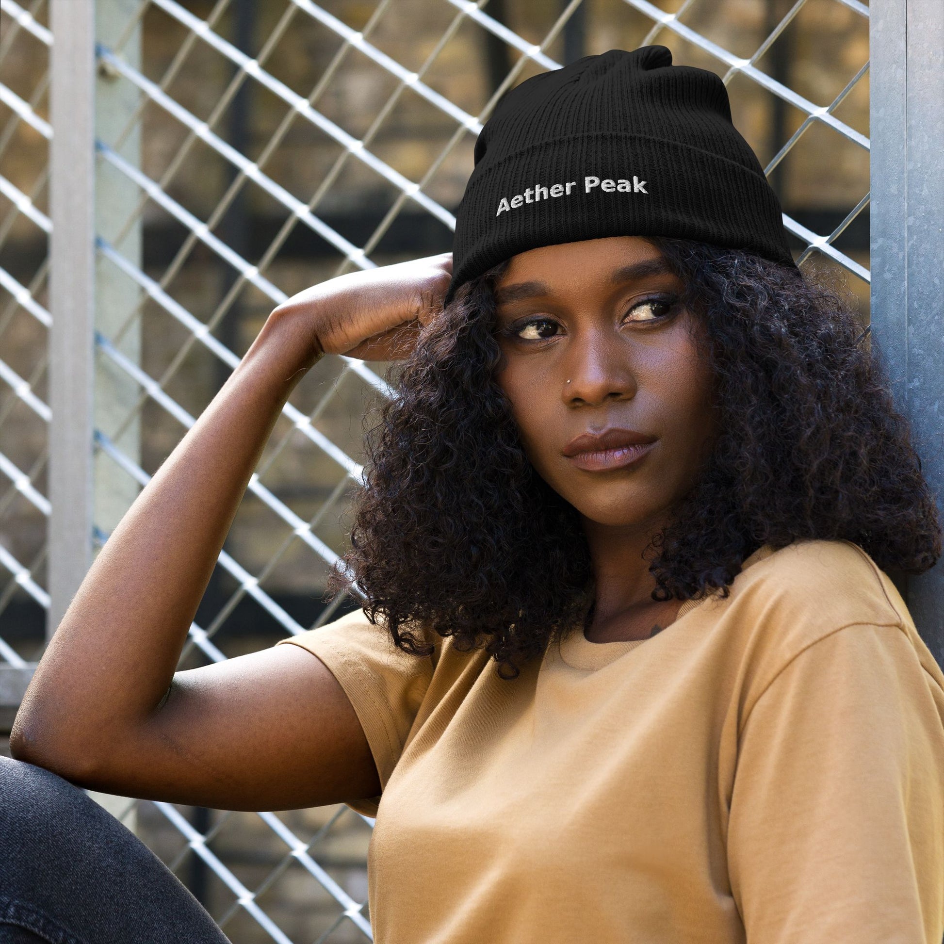 Woman wearing a black beanie with 'Aether Peak' text against a metal fence background