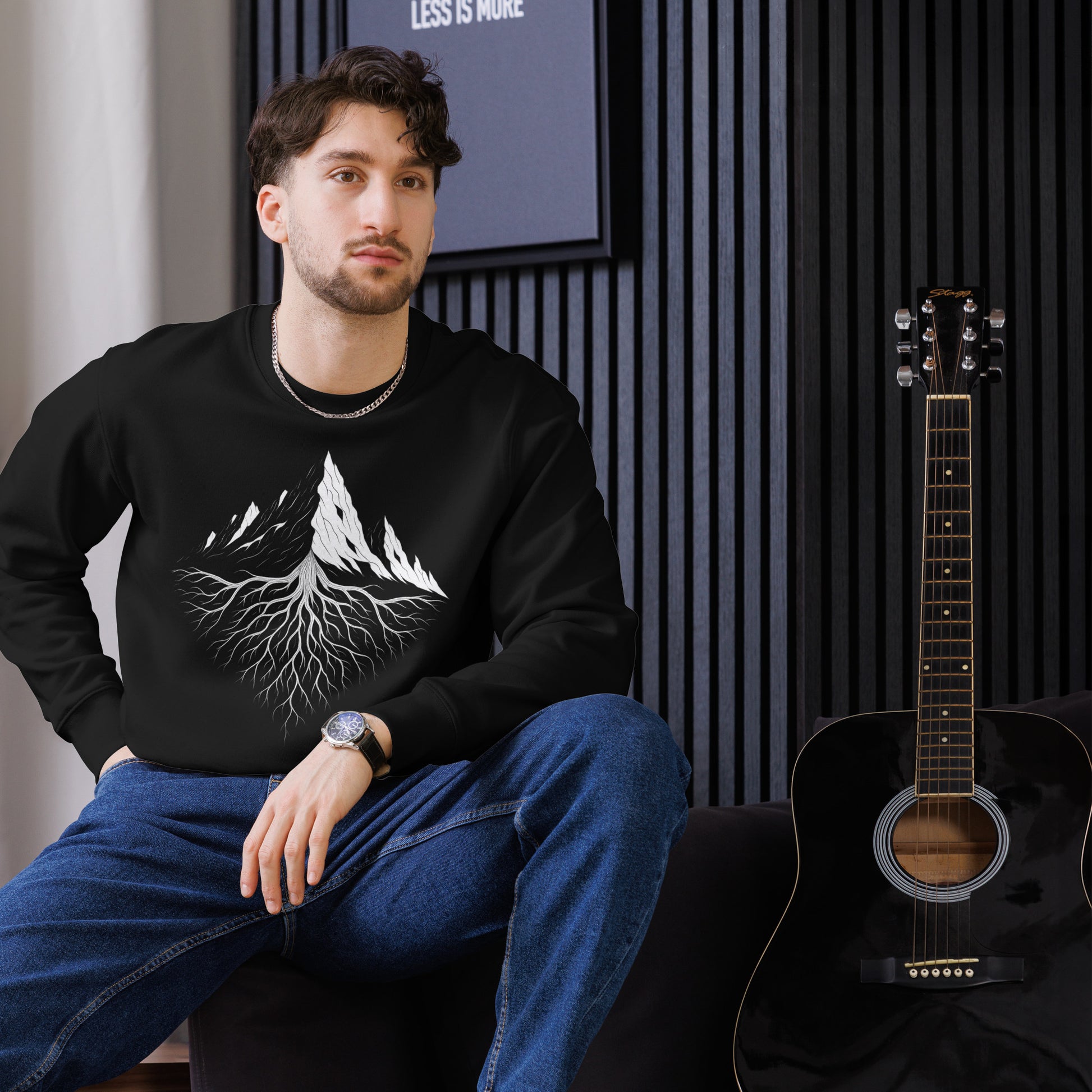 A man wearing the black Aether Peak sweatshirt with a white graphic design, sitting next to an acoustic guitar indoors.