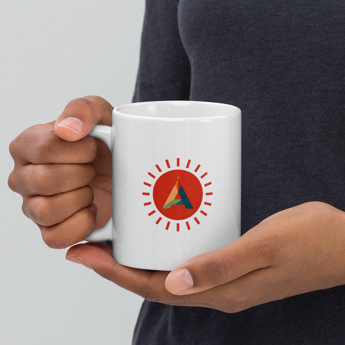 Hands holding a white mug with the Aether Peak sunburst logo, person wearing a dark top against a light background.
